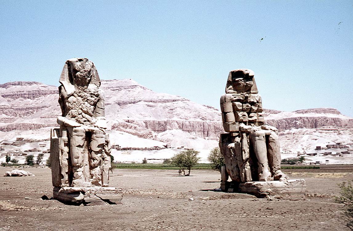 A group of stone statues in the desert with Colossi of Memnon in the background

Description automatically generated