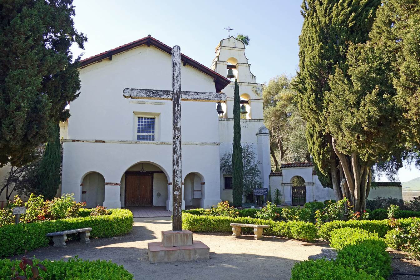 A white building with a cross in front of it with Mission San Juan Bautista in the background

Description automatically generated